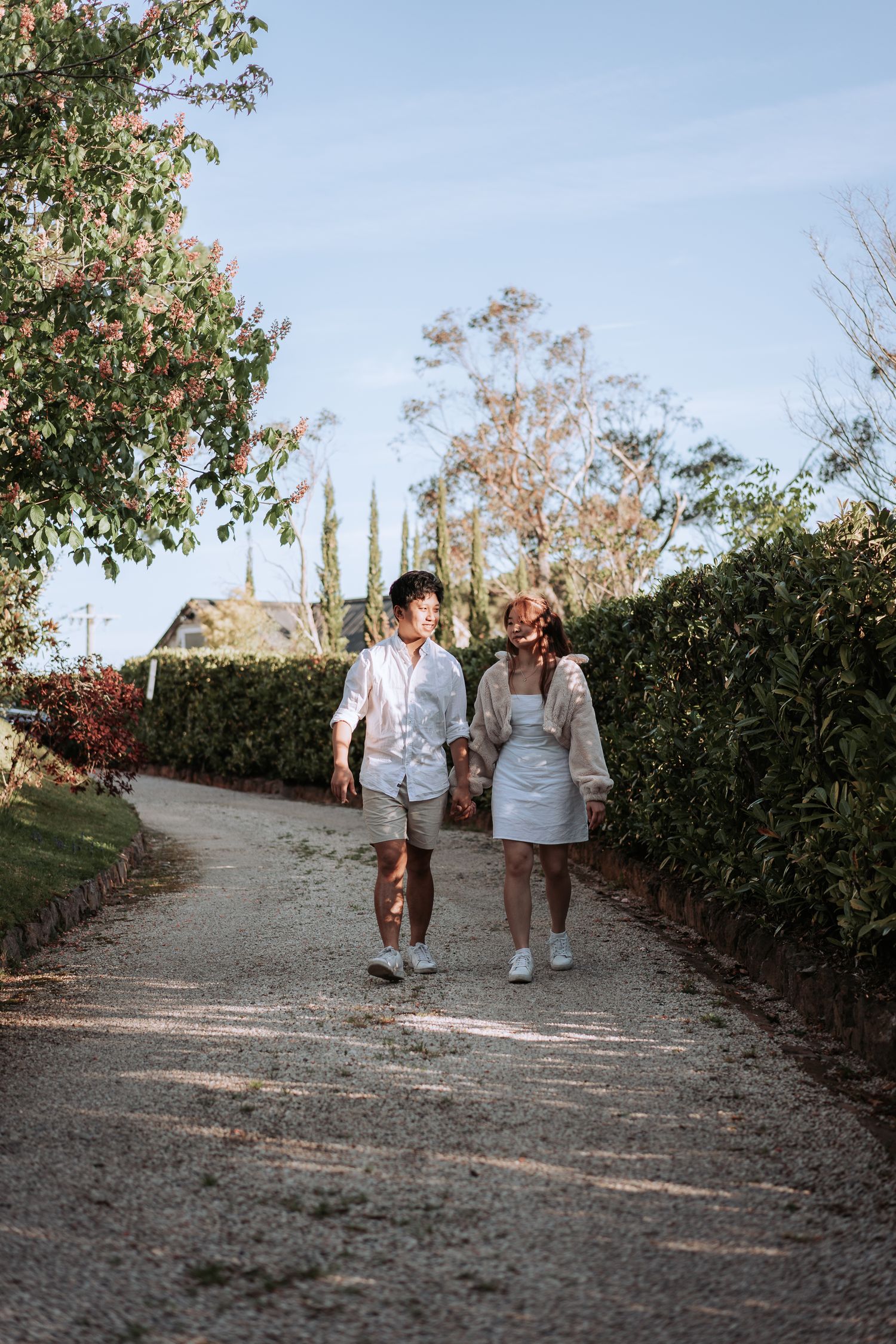 Two people in casual white outfits walking together on a curved garden path during sunset.