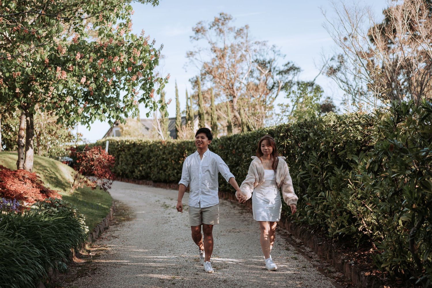 A couple holding hands while walking down a scenic garden path surrounded by lush greenery.