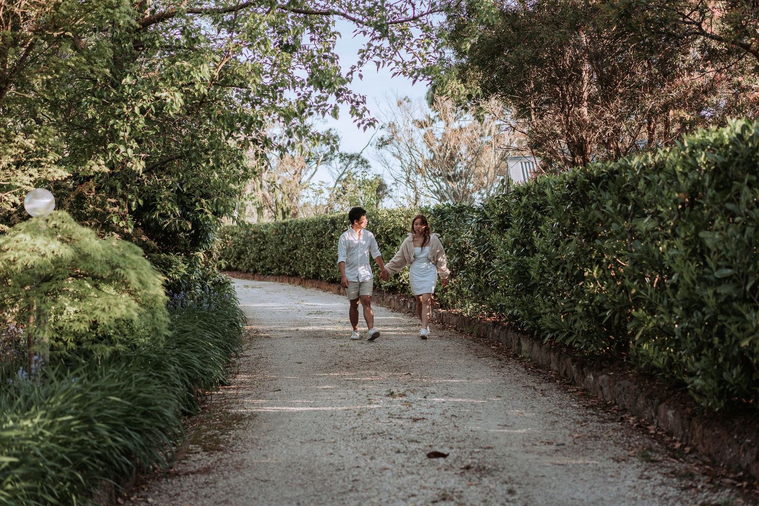Two people in white outfits walking together on a garden path with manicured hedges.