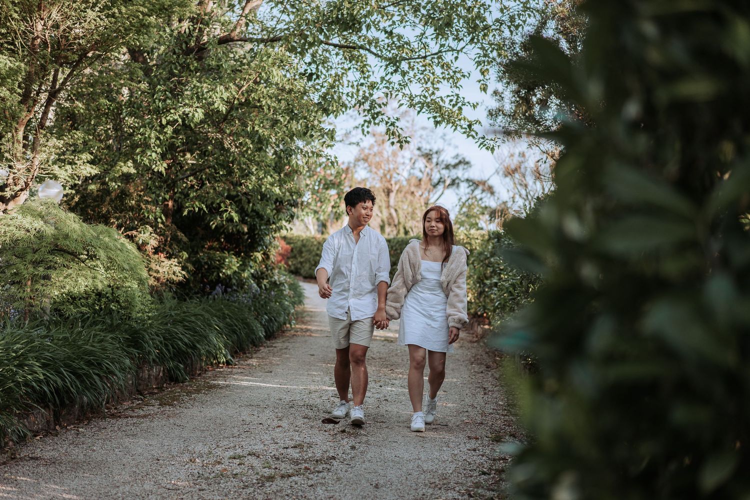 A couple in matching white outfits strolling down a tree-lined garden path.