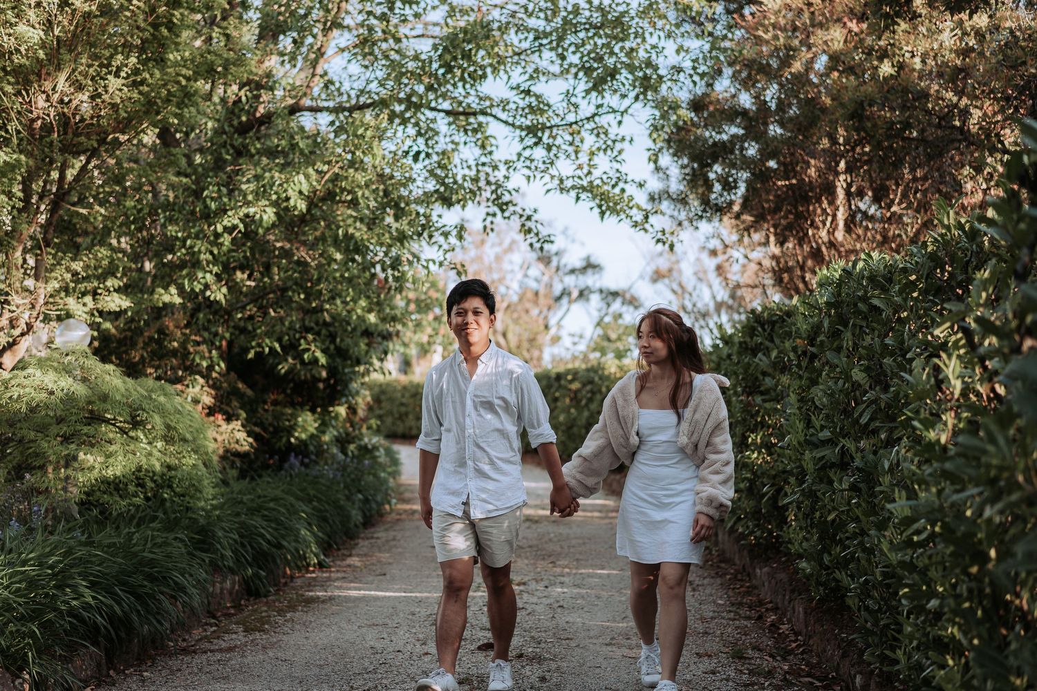 A couple in matching white outfits walk hand in hand down a tree-lined path on a sunny day.