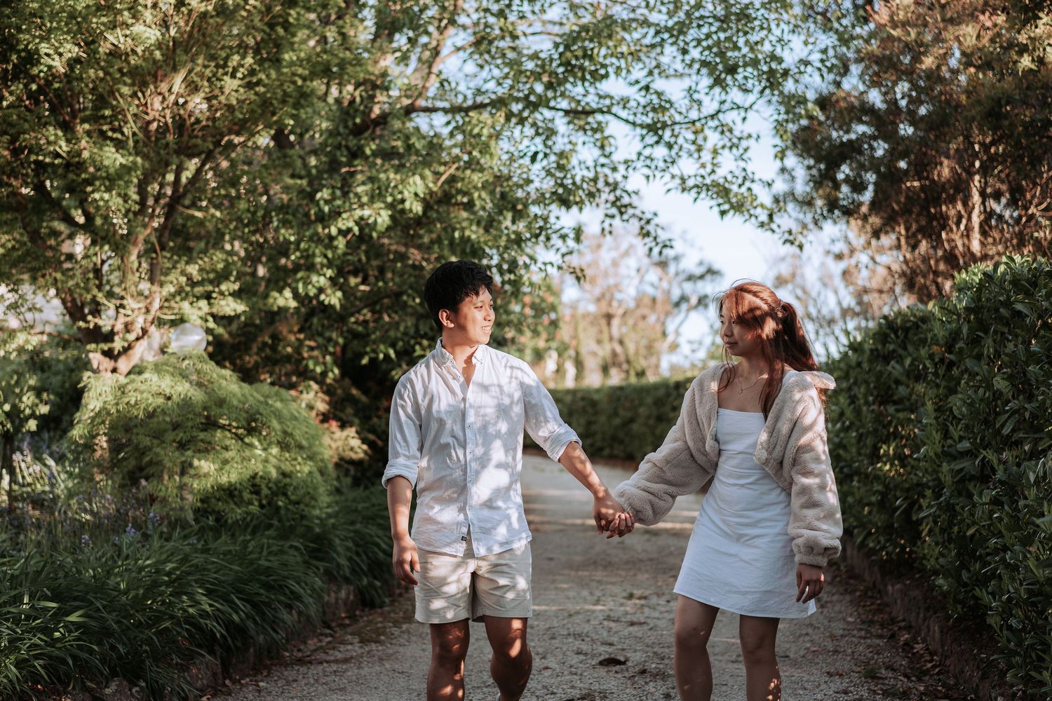 Two people in white clothing stroll together on a garden pathway while holding hands.