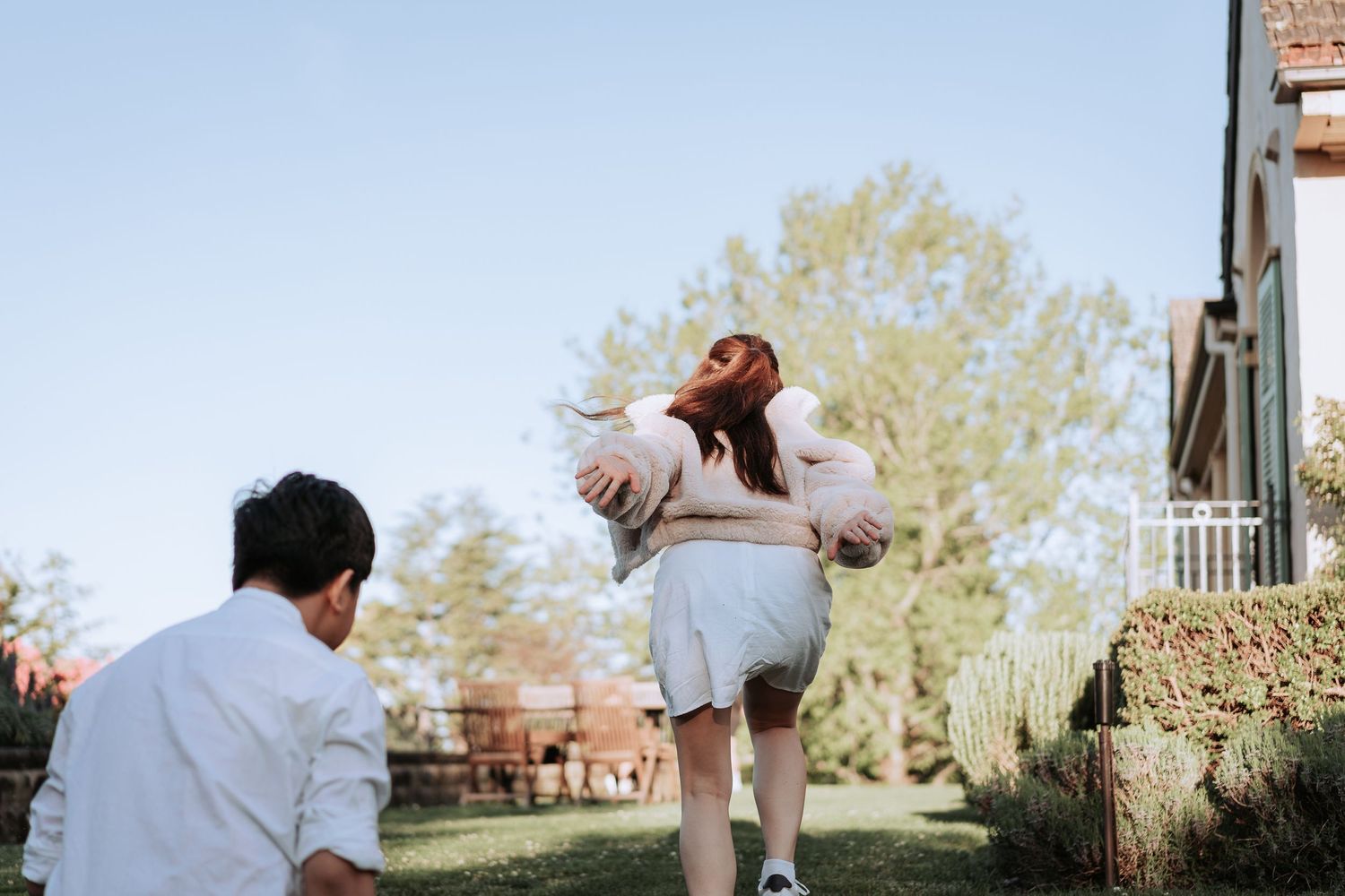 A person twirling joyfully in a white dress while another watches in a sunny garden setting.