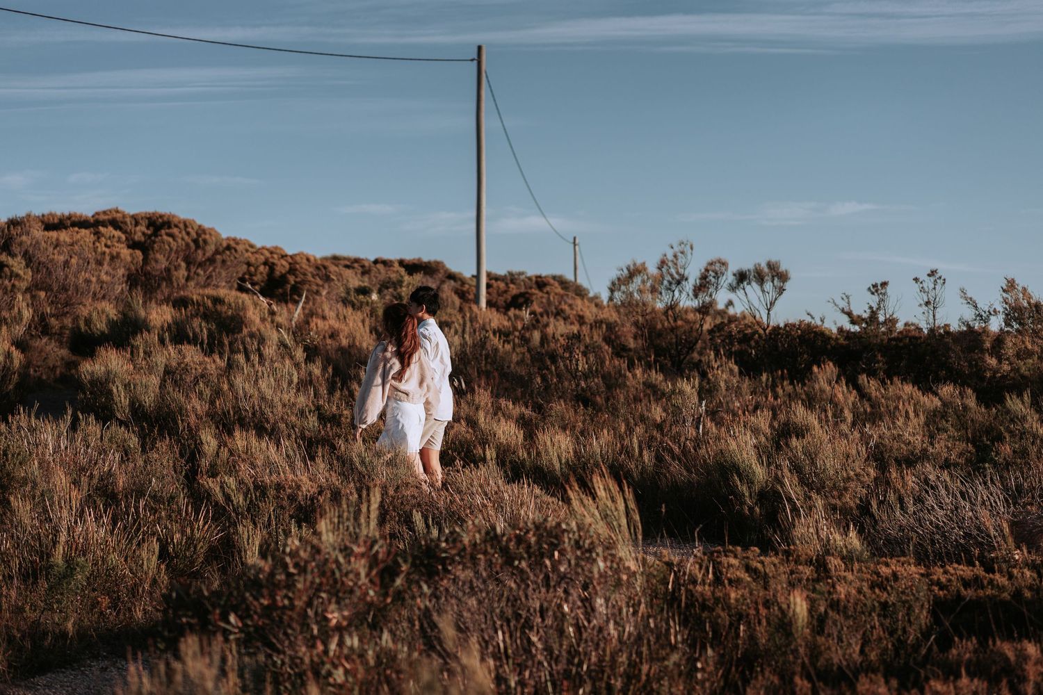 A figure in white stands on a rustic hillside path with utility poles.