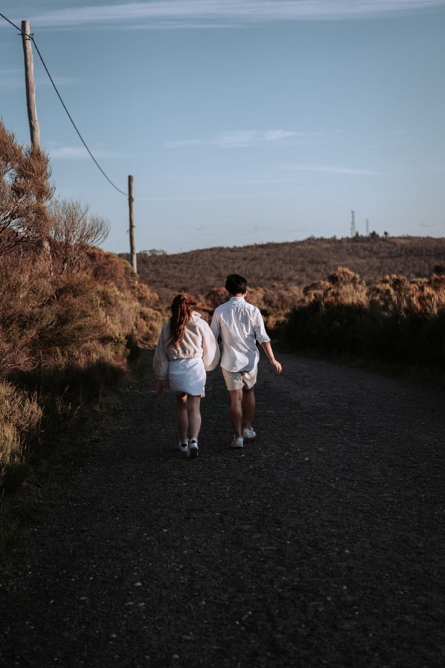 A couple walks away from camera on a dark mountain path under power lines at dusk.