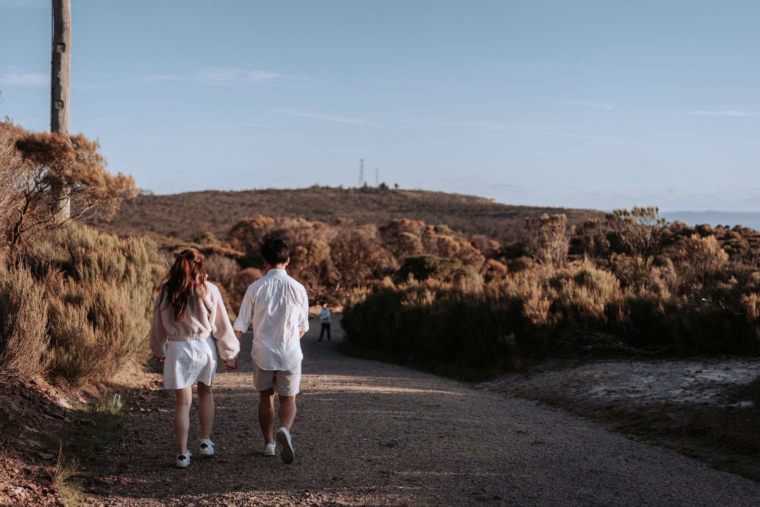 Couple in white clothing walking along a scenic dirt path surrounded by dry brush during golden hour.