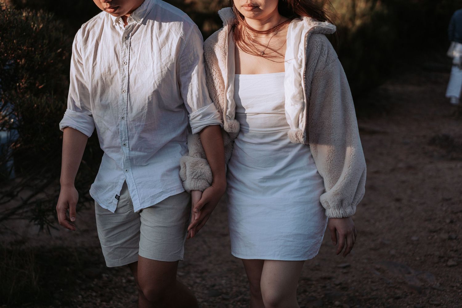 Two people in white outfits strolling together on a dirt path at dusk.