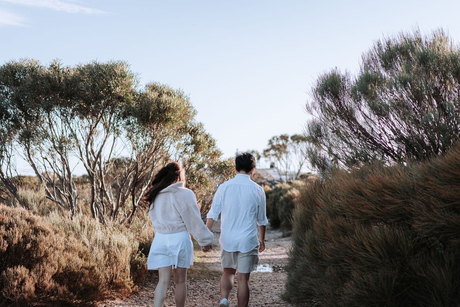 Couple in coordinated white outfits walking along a scenic mountain path under blue skies.