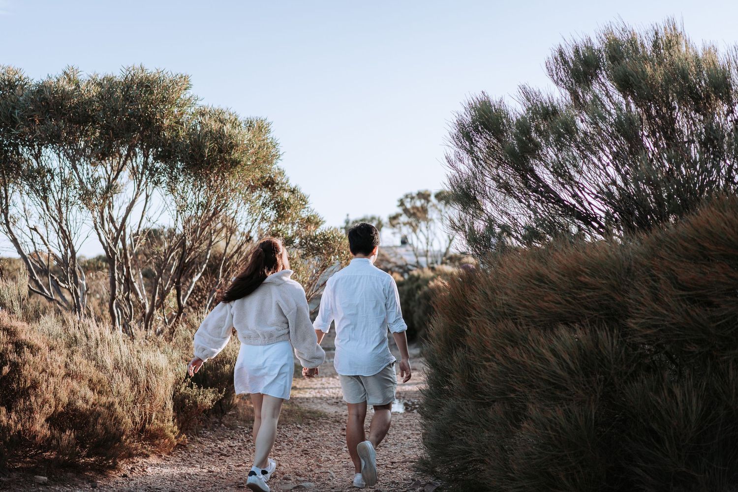 Couple in matching white outfits walking along a scenic hiking trail surrounded by shrubs.