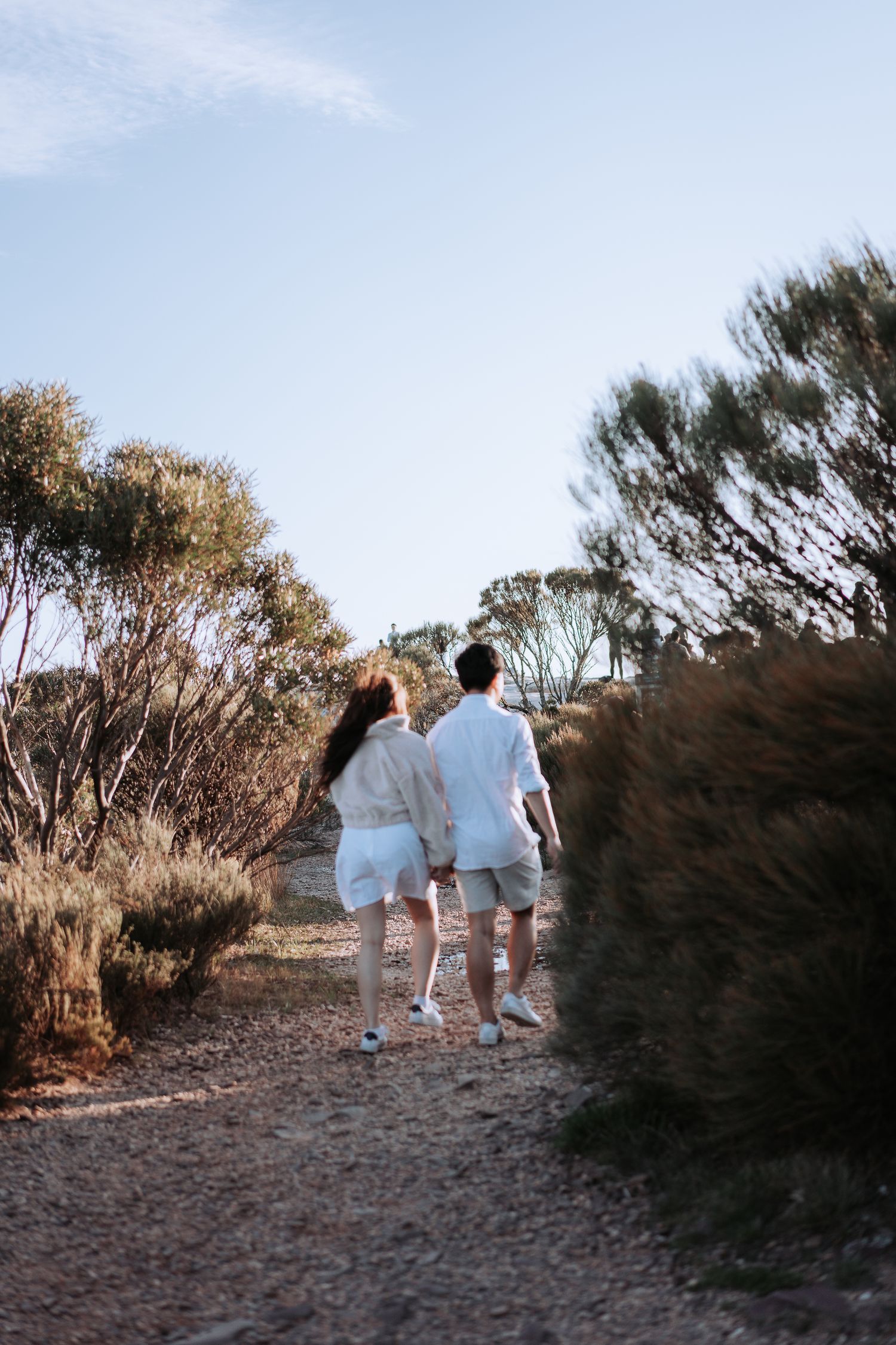 Two people in light clothing walking together on a nature trail lined with trees.