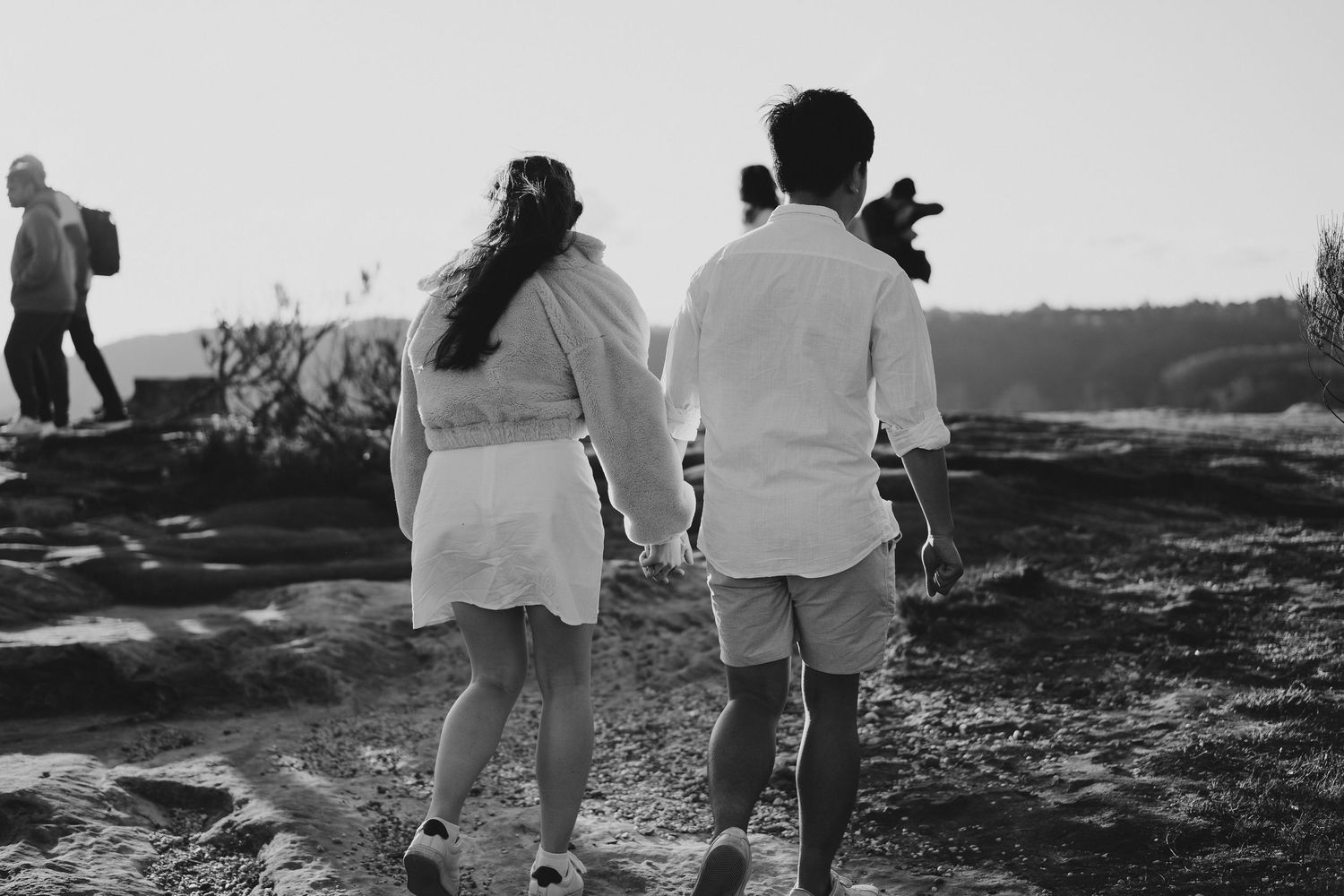 Black and white image of two people holding hands while walking on rocky terrain.