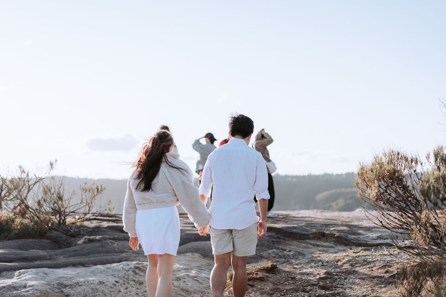 A couple in white outfits walking together on a rocky hiking trail overlooking mountain views.