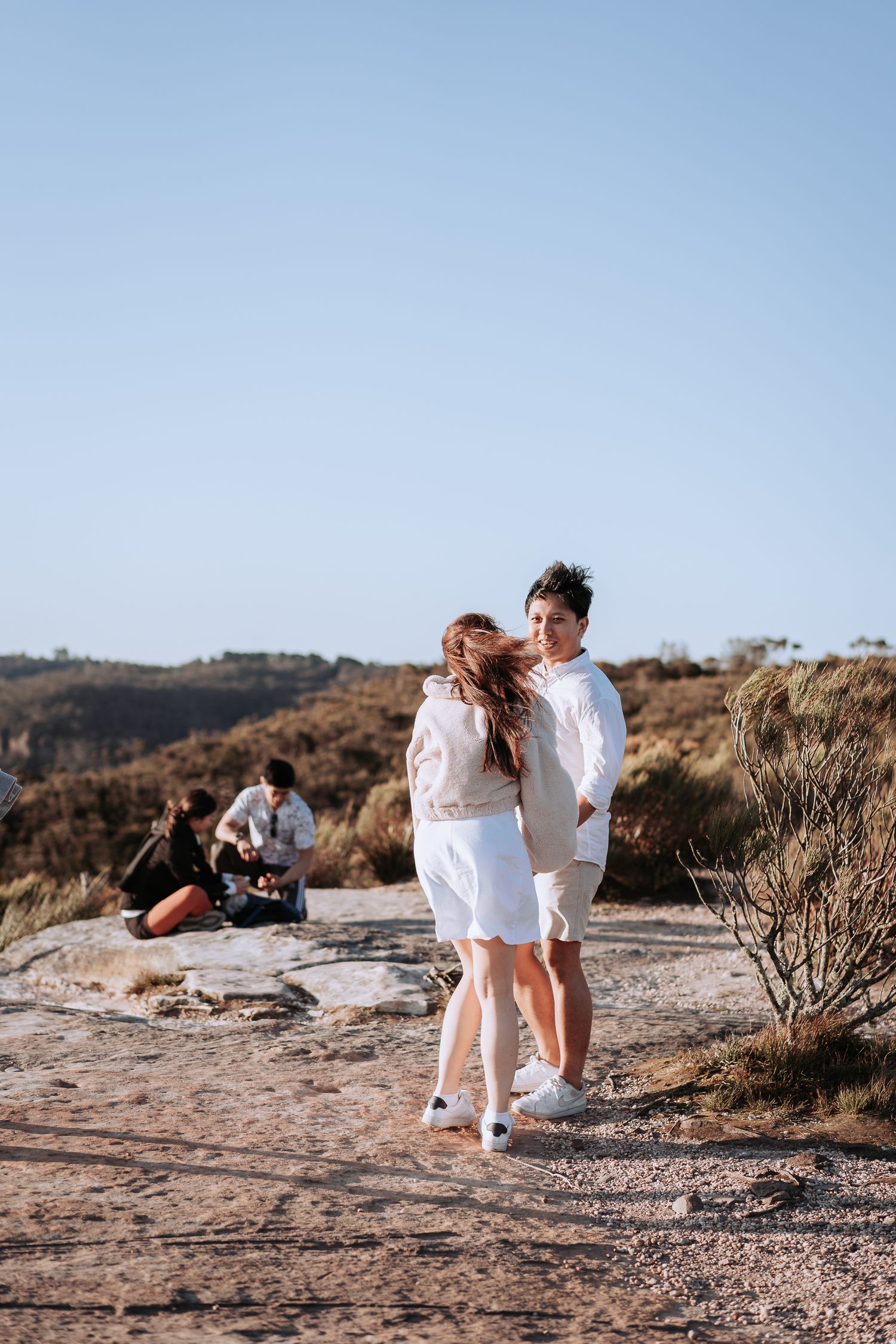 Couple in white outfits embracing on rocky mountain trail with scenic valley views.