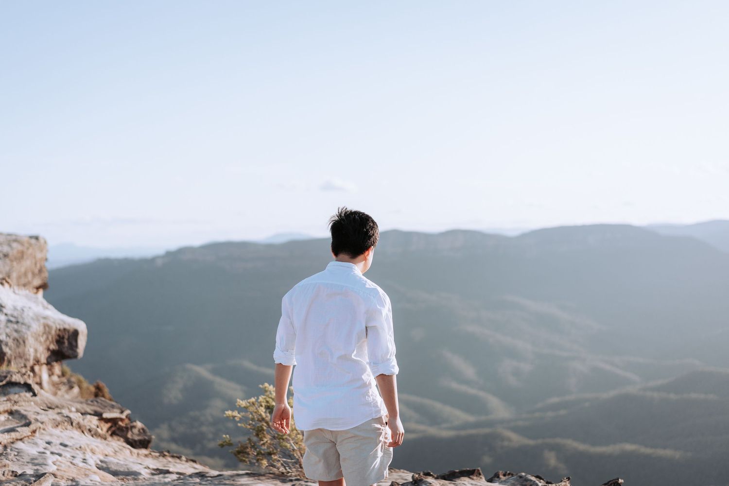 Person in white shirt standing on mountain overlook gazing at scenic valley landscape.
