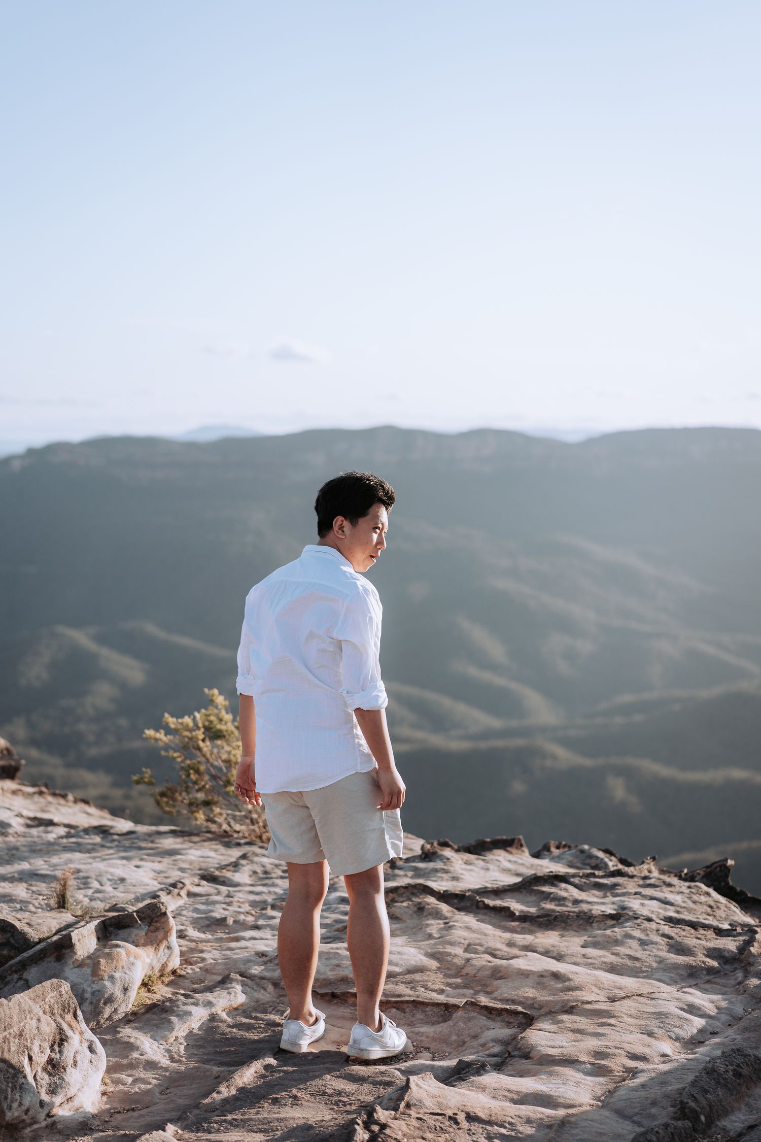 Person in white shirt and khaki shorts standing on rocky mountain ledge overlooking valley.