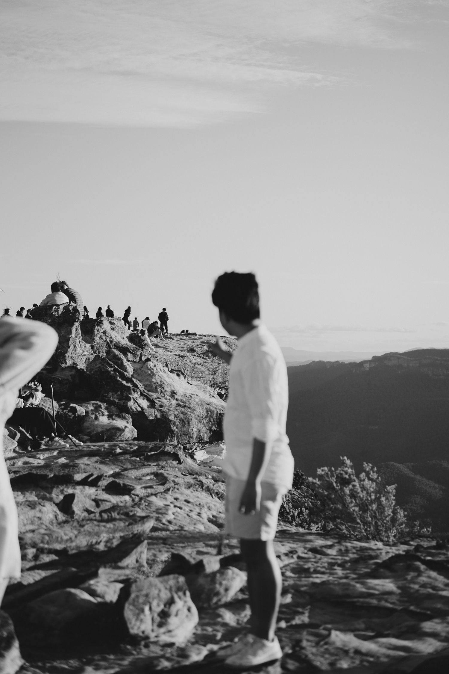 Black and white photo of someone on mountain summit with crowd of hikers visible in background.