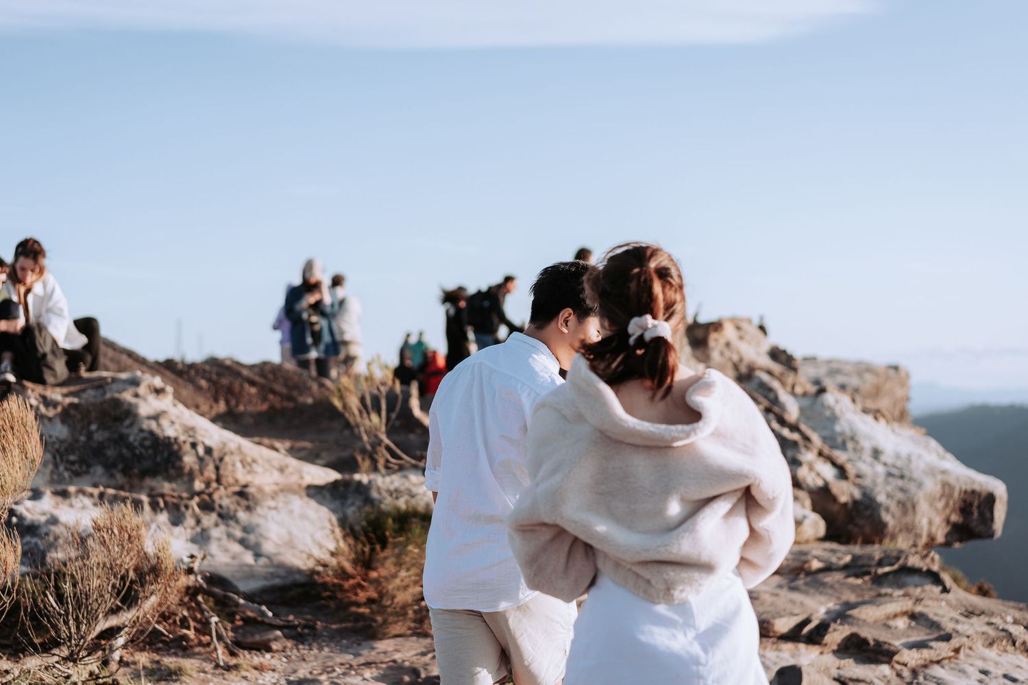 Couple in white embracing on rocky mountain overlook with other hikers visible in background.