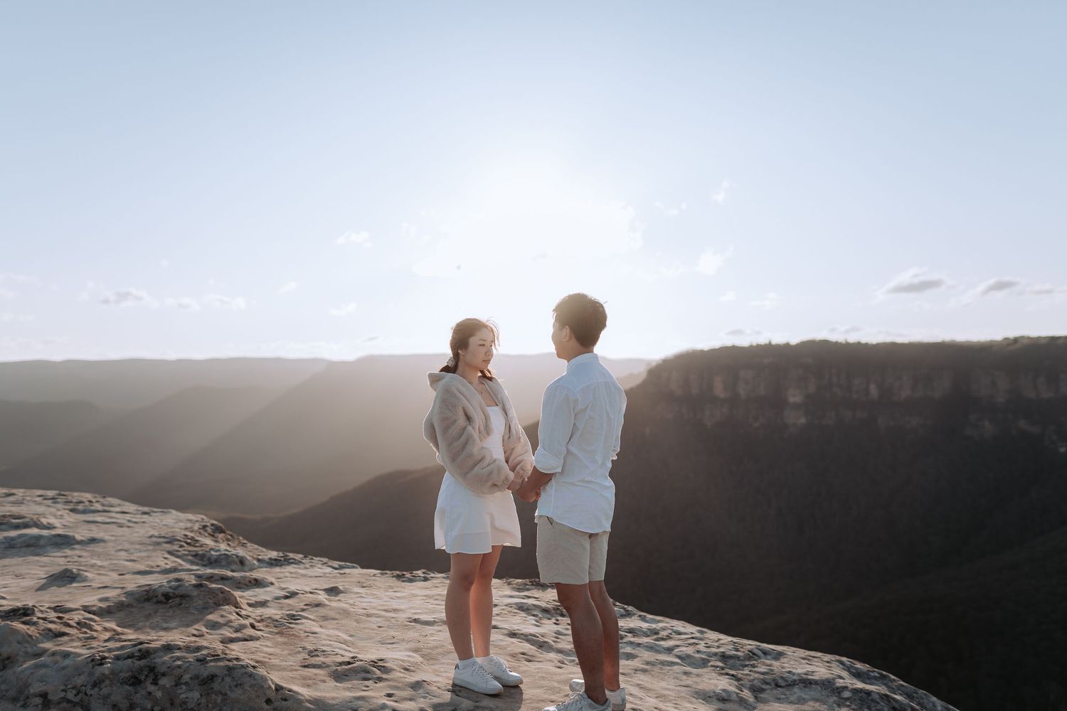 A couple stands together at the edge of a cliff during golden hour enjoying scenic mountain views.