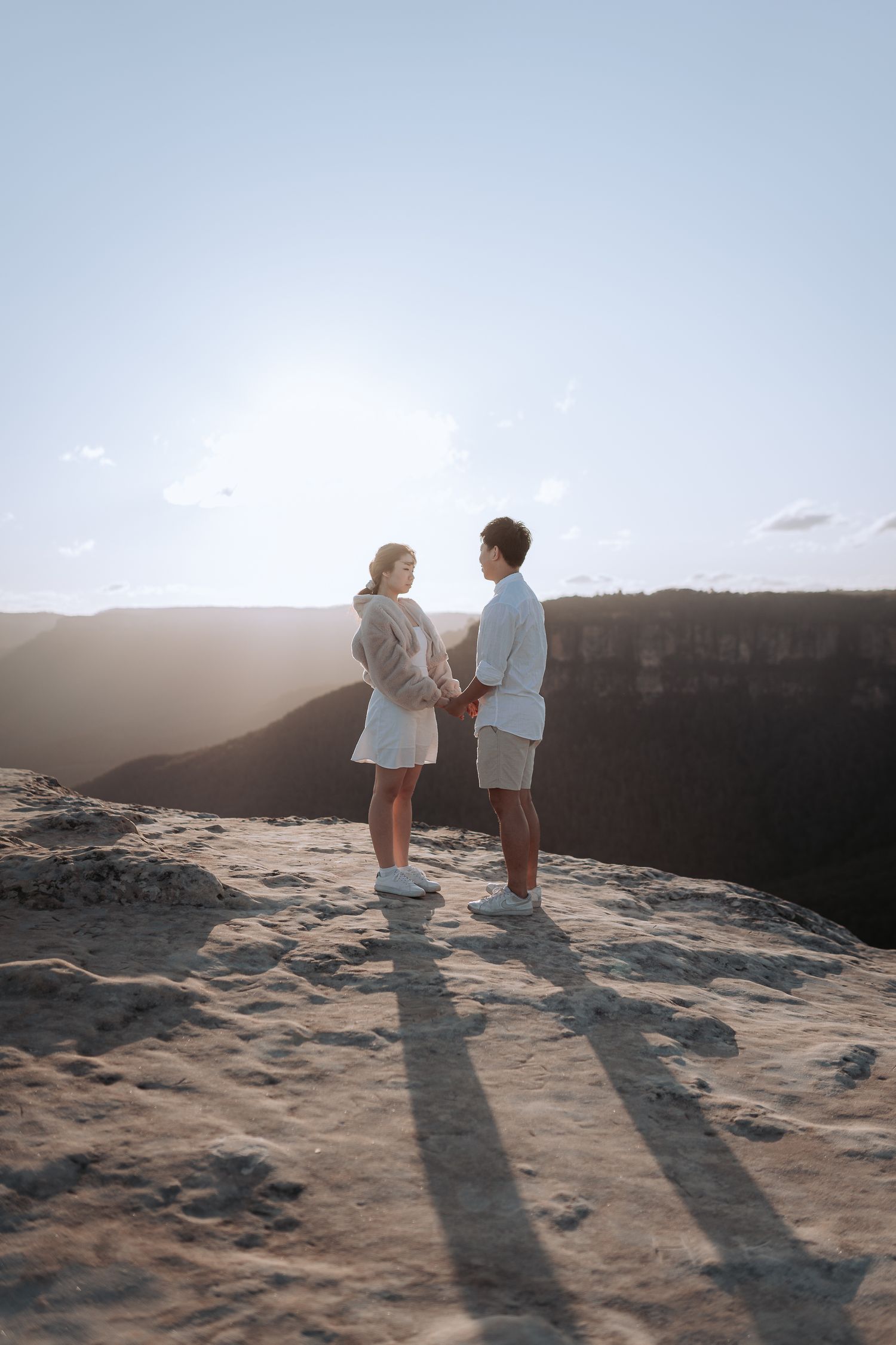 Two people cast long shadows while standing on a rocky cliff edge at sunset.