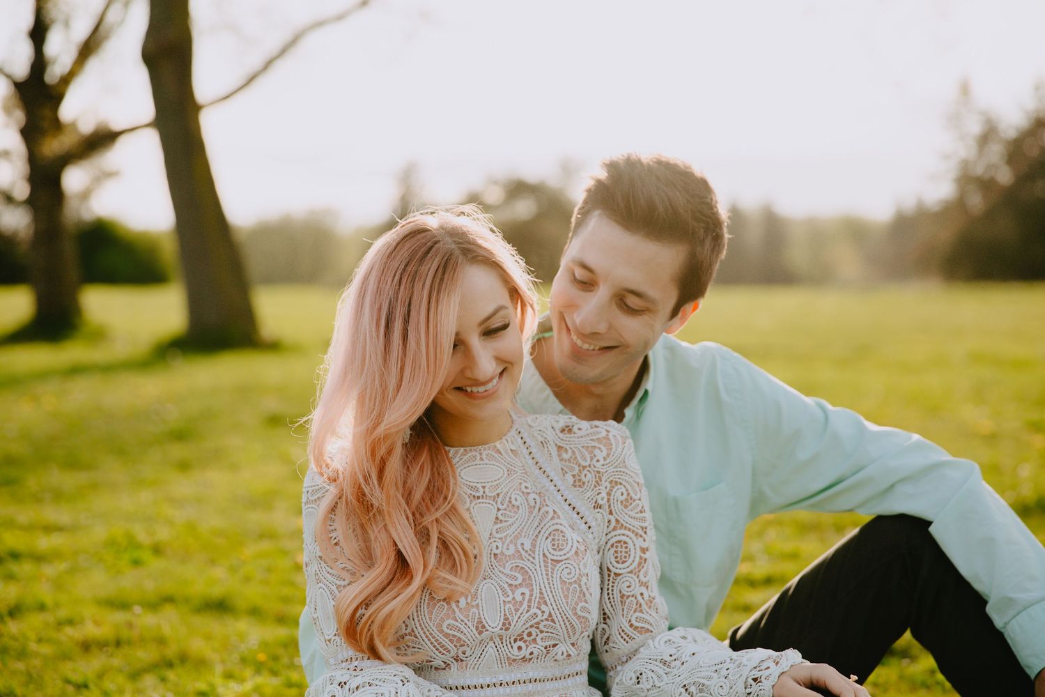 Two people share an intimate moment while sitting together on grass in a sunlit park setting on a warm afternoon.