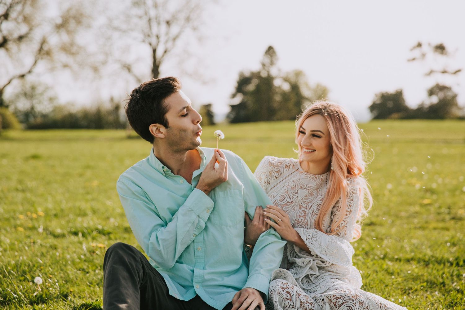 A couple sits together in a grassy meadow on a sunny afternoon sharing a playful moment.