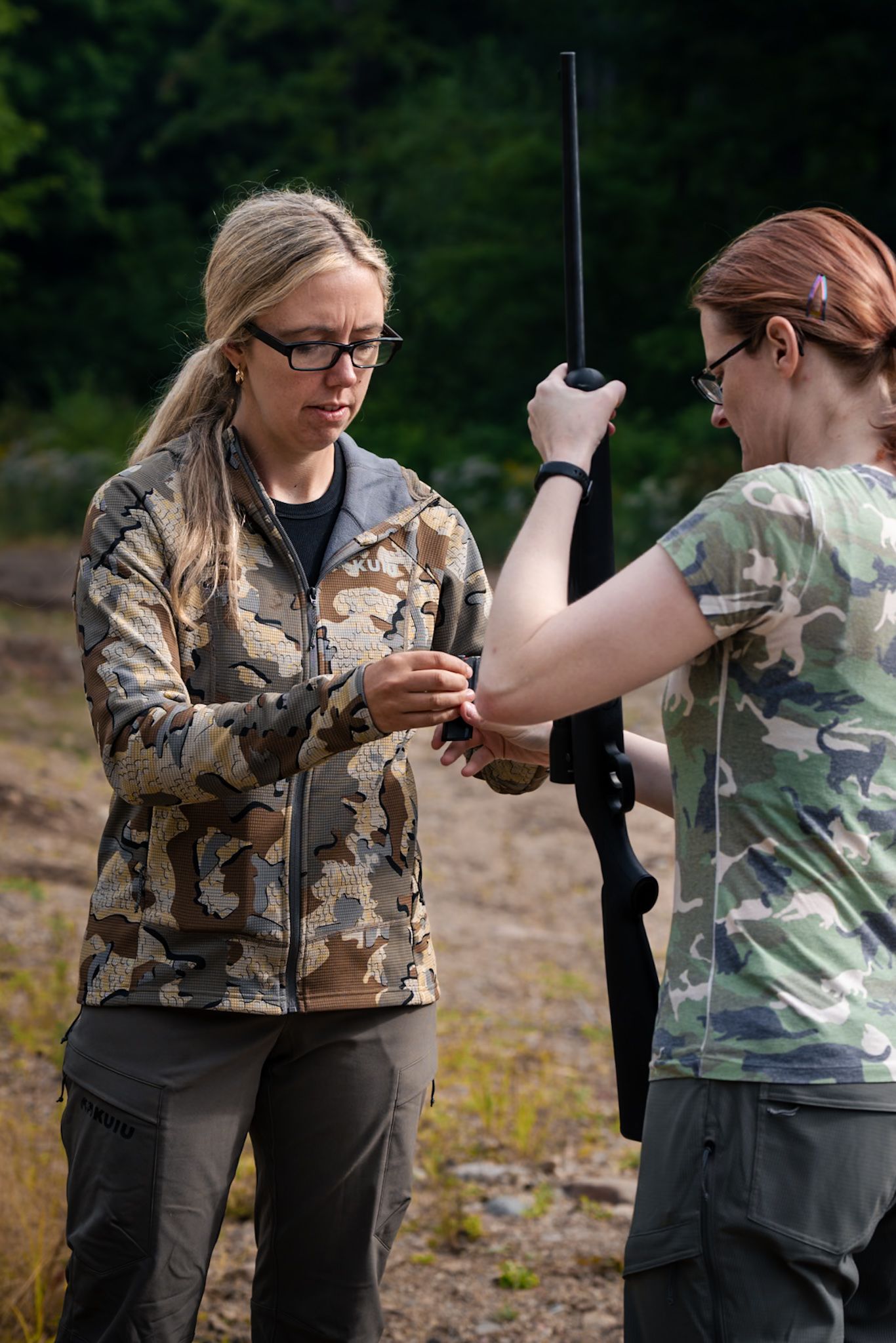 Two people in camouflage gear inspect firearms and equipment during an outdoor training session.
