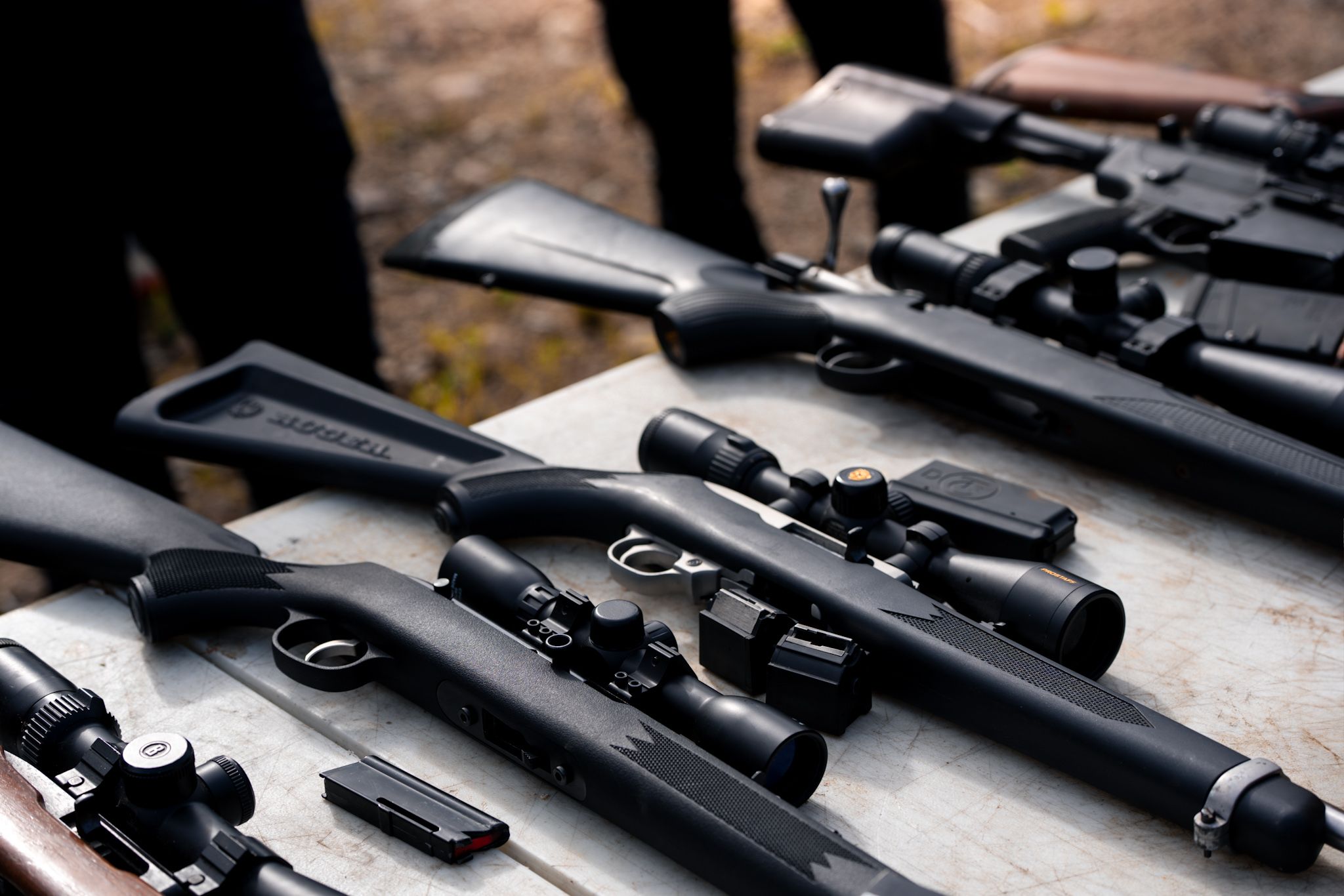 Close up view of multiple black rifles with mounted scopes lined up on a wooden surface.