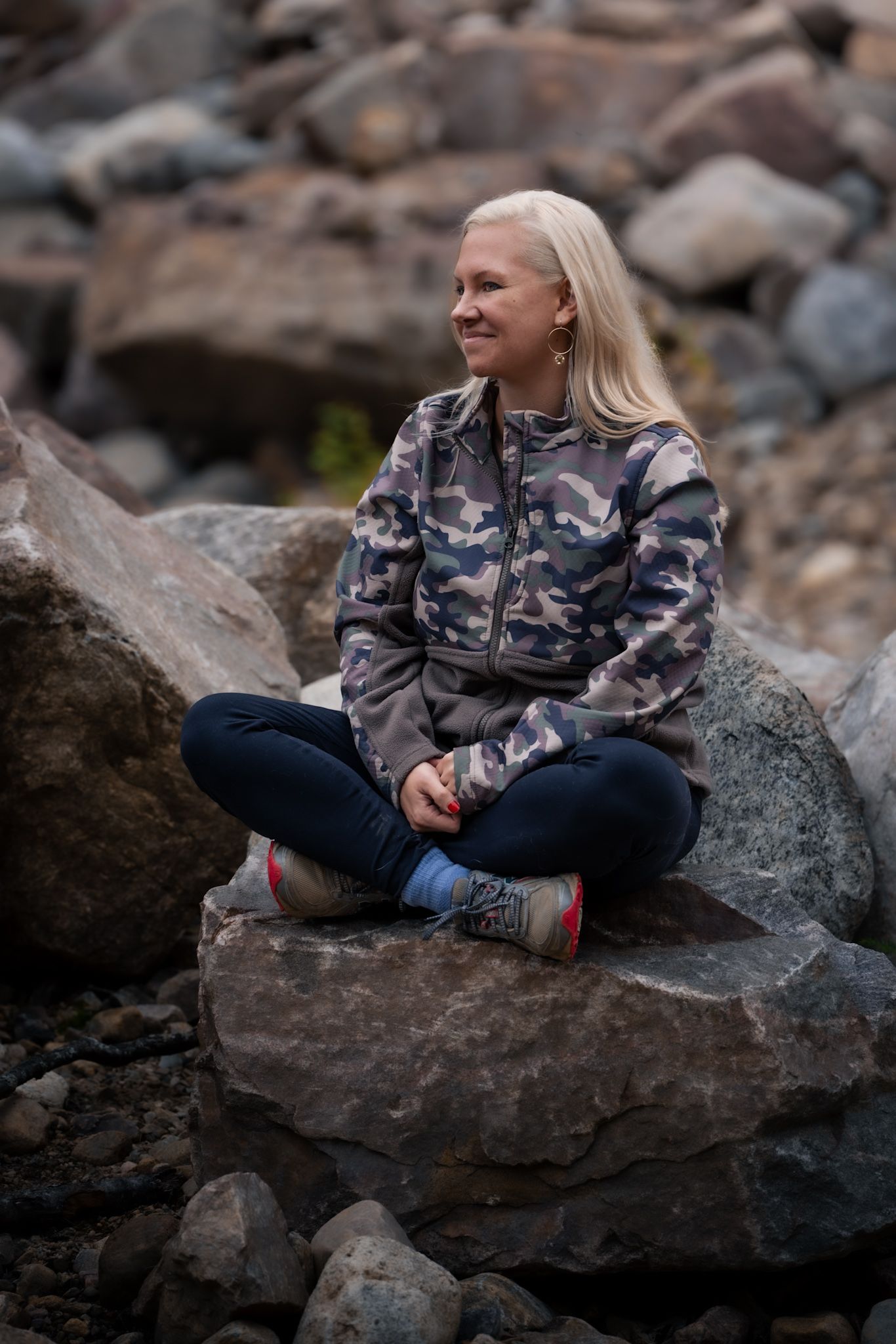 Person in camo jacket sits cross-legged on large rock formation with stones and boulders in background.