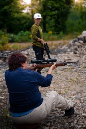 Person practices target shooting with a scoped rifle in rocky outdoor terrain while receiving instruction.