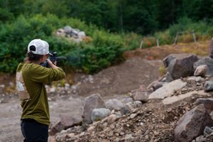 Person wearing a baseball cap and olive jacket takes photos on rocky terrain surrounded by green foliage.