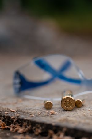 Empty bullet shell casings lie on concrete with protective blue safety glasses blurred in the background.