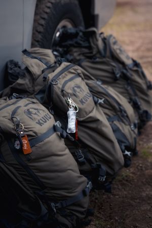 Row of brown hiking backpacks with gear attachments and carabiners lined up on dirt ground outdoors.