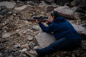 A person in a blue jacket lies on rocky terrain while aiming a scoped rifle.