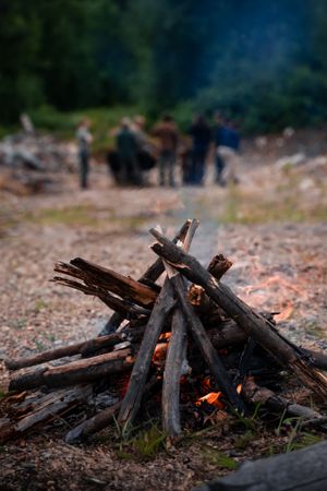 A campfire with stacked wooden logs begins to ignite while a group gathers in the blurred background.