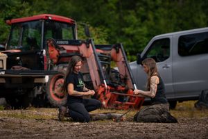 Two people sit on rocks having a conversation with a red tractor and silver vehicle in the background.