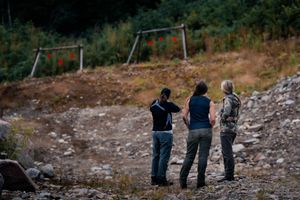 Three people stand together at a shooting range with an old swing set in the background on rugged terrain.