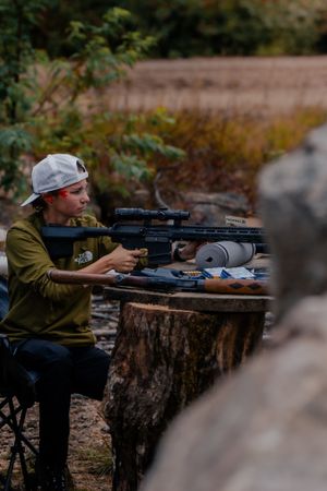 A shooter in an olive shirt and gray cap aims a scoped rifle while seated at a rustic outdoor shooting station.