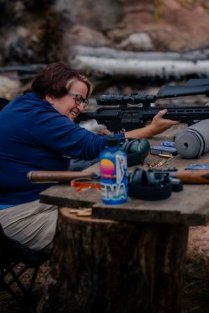 A shooter in a blue shirt practices with a scoped rifle at an outdoor range setup on a tree stump.