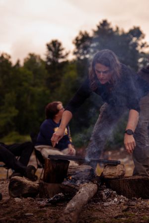Campers tend to a smoking campfire at sunset in the wilderness while relaxing outdoors.