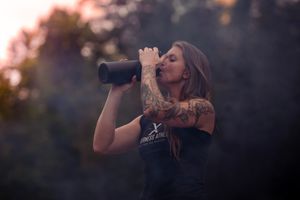 A person in a black tank top drinks from a water bottle at sunset with a blurred bokeh background.