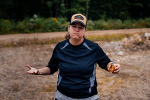 A person on a dirt path wearing a navy blue shirt and trucker hat gestures with both hands.
