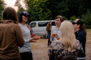 A group of people gather outdoors in conversation, enjoying drinks on a warm day with a parked vehicle in the background.