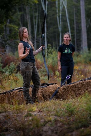 Two archers with bows stand on a wooded hillside during outdoor target practice in natural terrain.