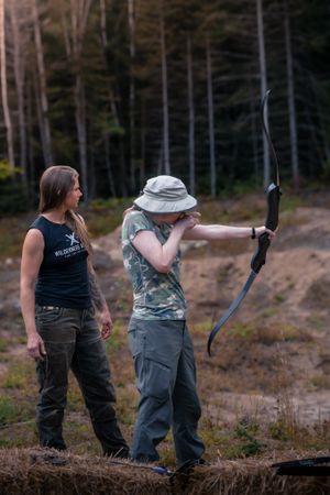 Two people practicing archery outdoors with one taking aim using a bow while standing on rough terrain near a forest.