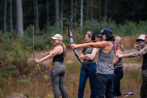 Several people practice archery outdoors in a field surrounded by trees.
