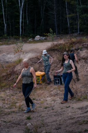 A group walks along a dirt trail through wooded terrain carrying supplies and equipment.