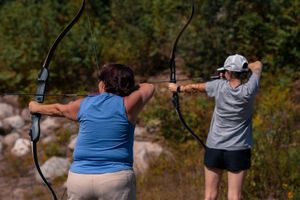 Two archers practicing with compound bows at an outdoor range on a sunny day.