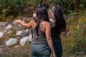 Two people with dark hair wearing tank tops stand close together outdoors against a blurred nature background.