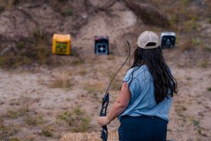 Archer wearing baseball cap and denim vest aims compound bow at outdoor target range on desert terrain.