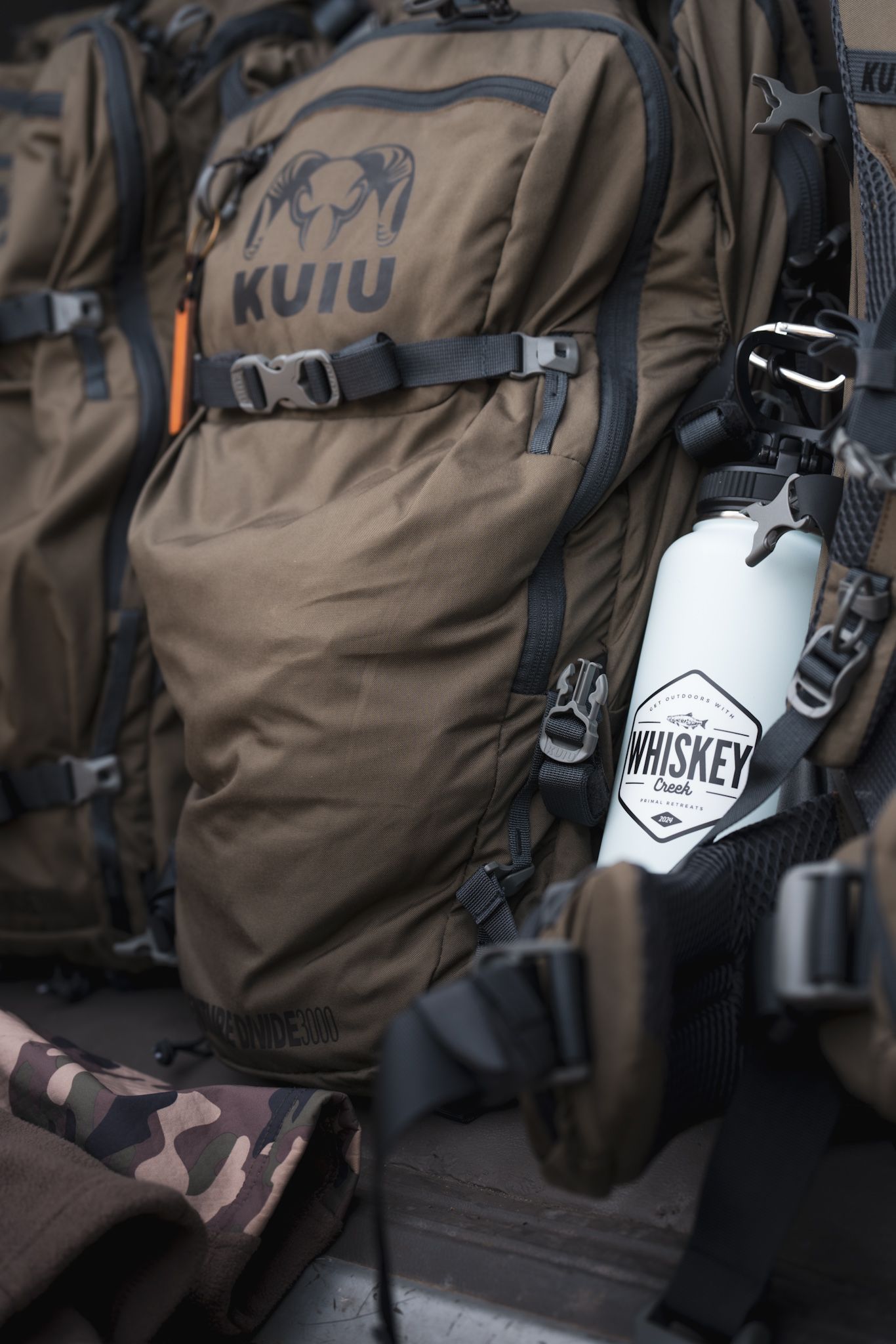 Close-up of brown Kuiu backpack with whiskey flask attachment and camo pattern gear.